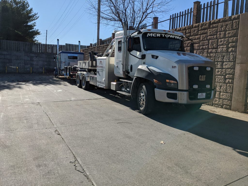 MCR Towing Cat CT660 wrecker on a concrete lot beside a stone wall, blue semi behind