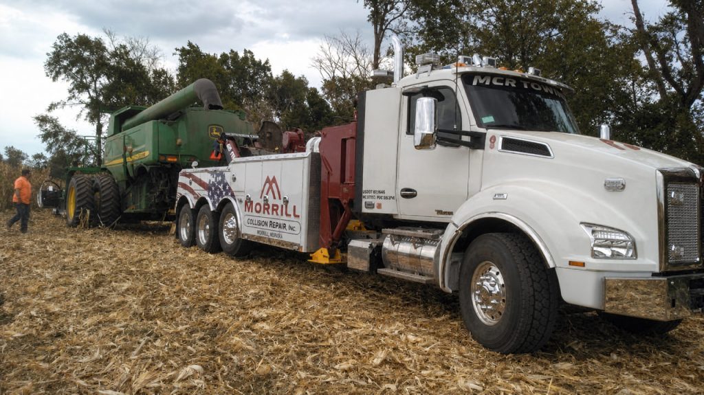 MCR Towing heavy wrecker in a harvested cornfield beside a John Deere combine and crew member