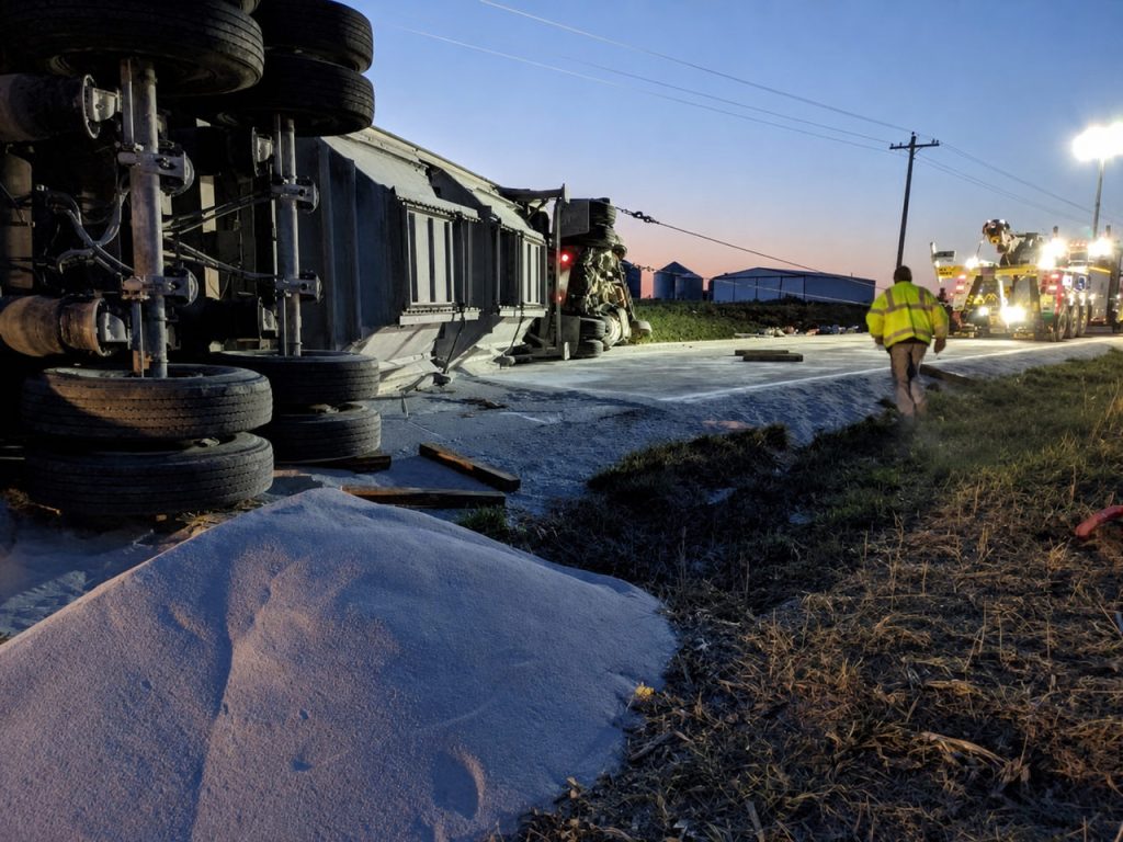 Semi trailer on its side with a spilled load at dusk, heavy wreckers lit beyond